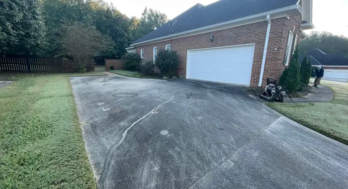 A concrete driveway darkened with algae and dirt in front of a brick house, a pressure washer standing at its edge