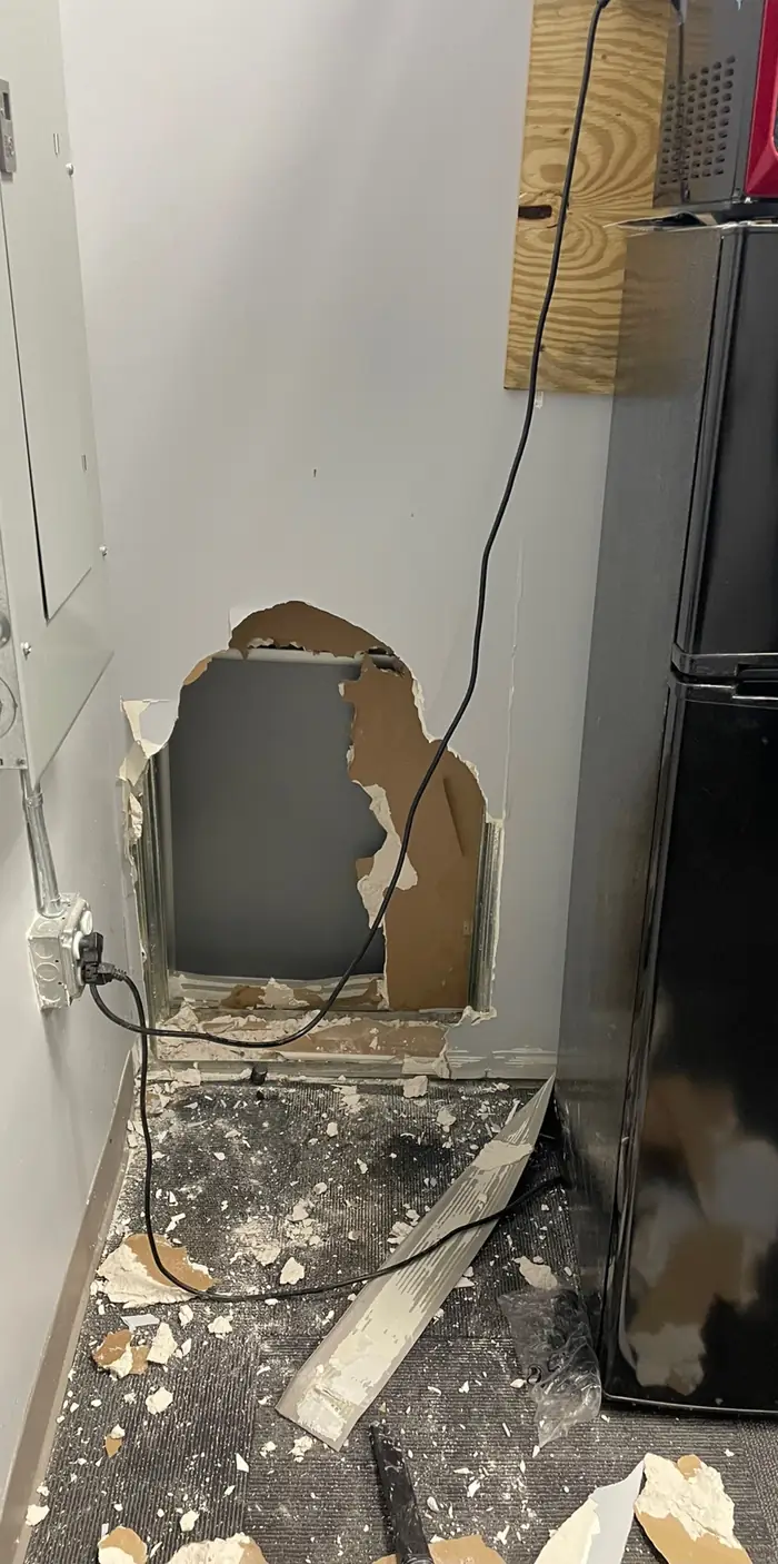 A broken hole in a drywall wall beside a refrigerator, pieces of board and dust on the floor below