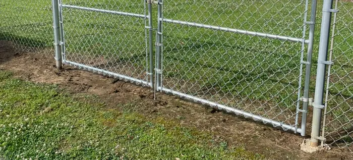 A closed double chain link drive gate at the end of a fence run, its posts standing in freshly turned dirt