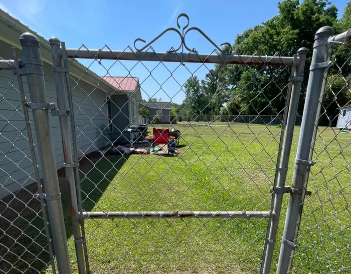 A chain link gate with a scrolled metal arch across the top, hung between galvanized posts, a mown yard behind it