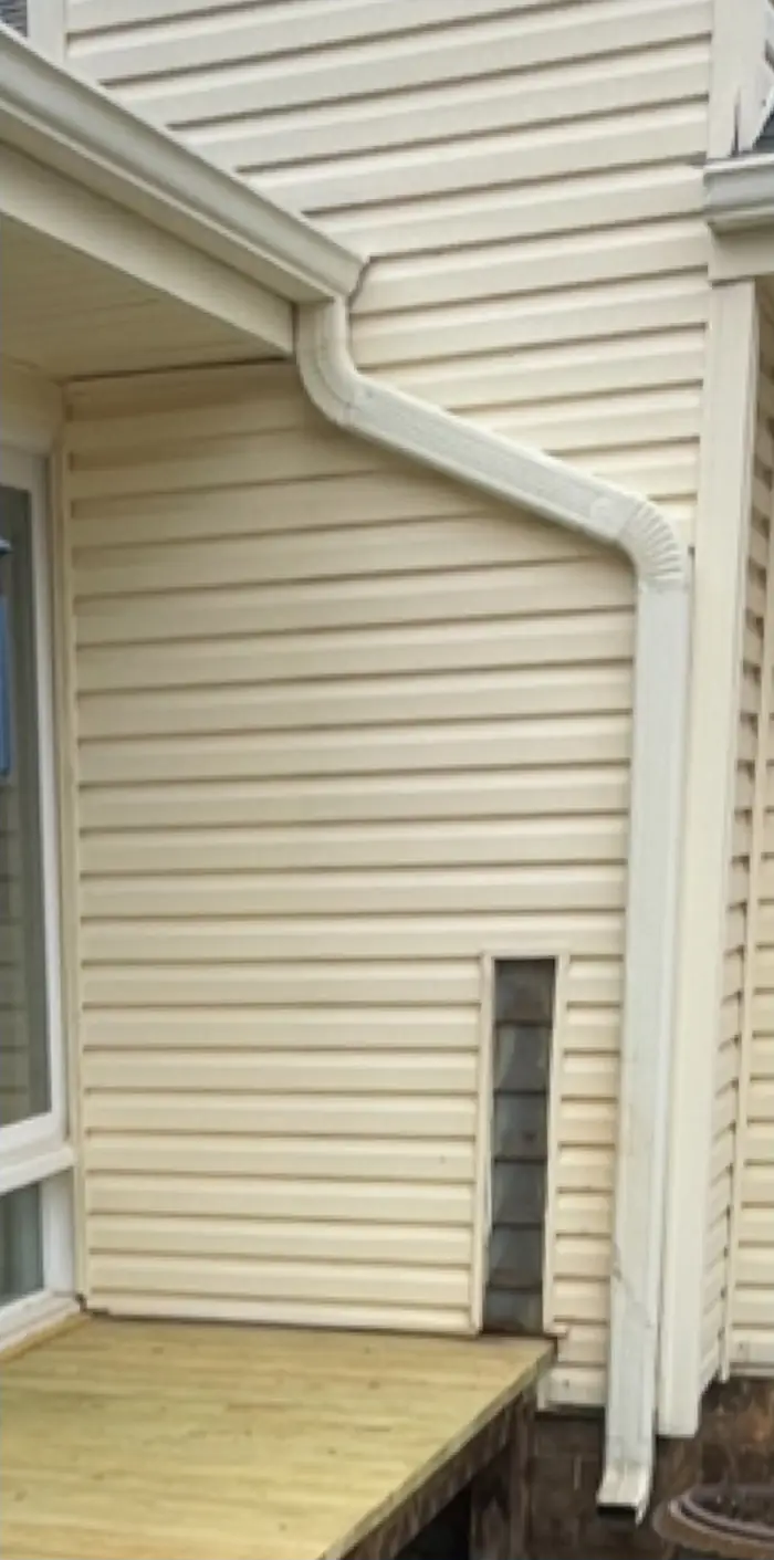 A white gutter and downspout run down an outside corner of cream vinyl siding, above a wood deck