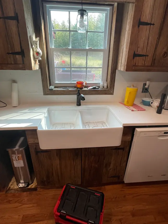 A white apron front sink set into a counter between dark stained cabinets, under a window with a pendant light