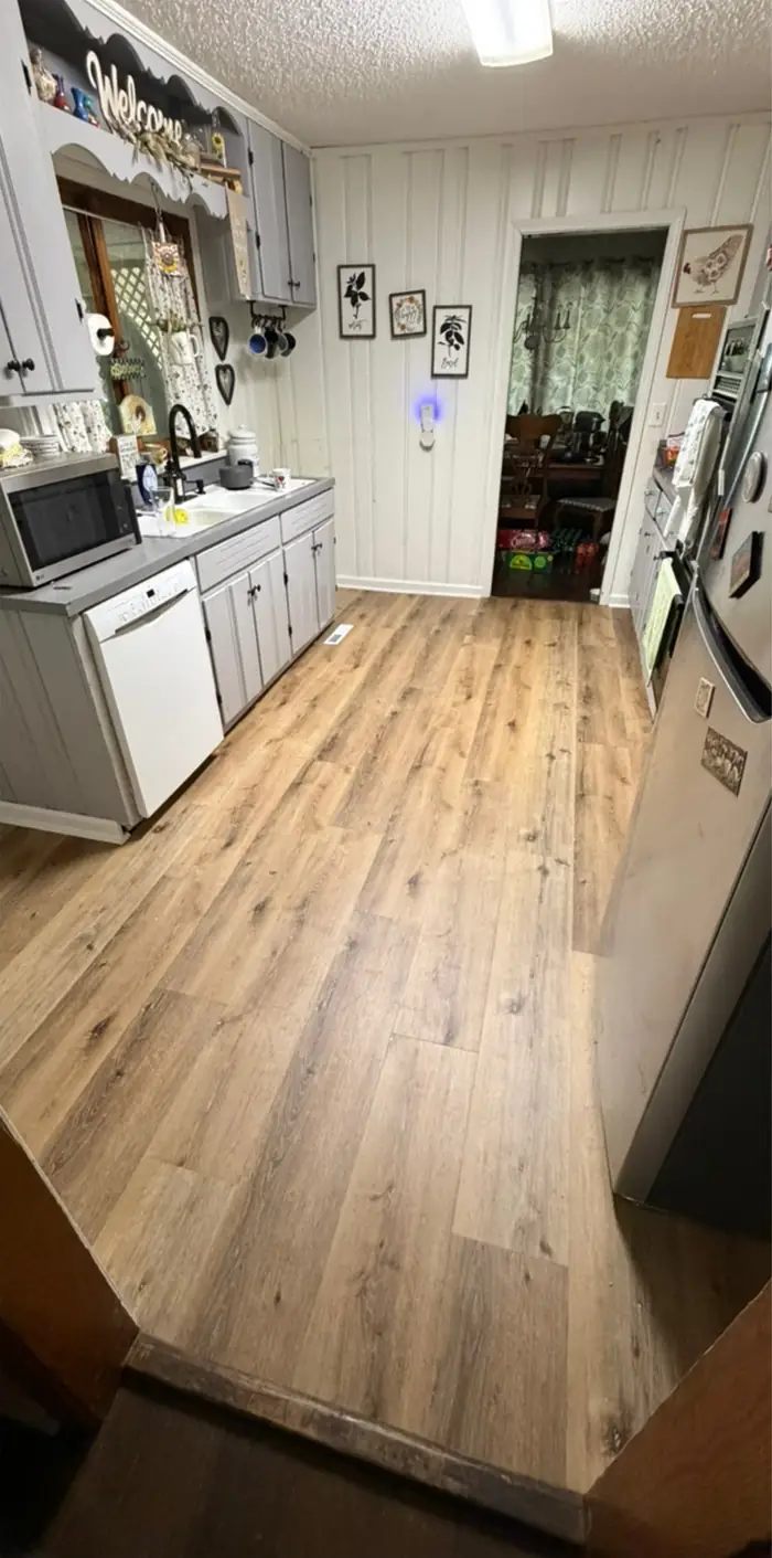 A kitchen floored in wide wood-look planks running to a dining room doorway, meeting the older floor at a threshold