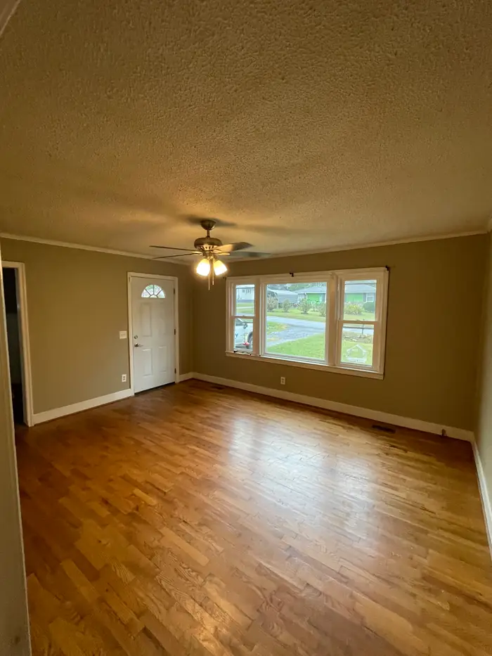 An empty room painted warm tan with white trim, a hardwood floor, a ceiling fan and two front windows