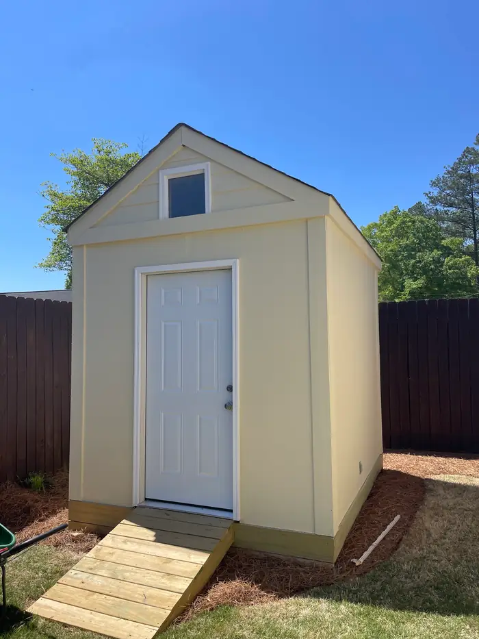 A storage building with a white door and a gable vent, a new wood ramp up to the threshold, against a wood privacy fence