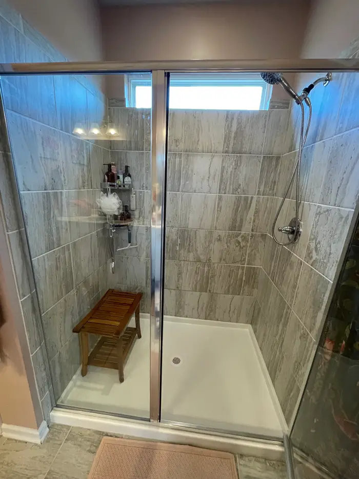 A tiled walk in shower with a wood bench set on the white shower base behind sliding glass doors