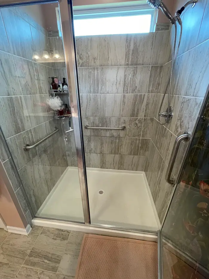 A walk in shower tiled in gray wood-look tile, with sliding glass doors and two stainless grab bars